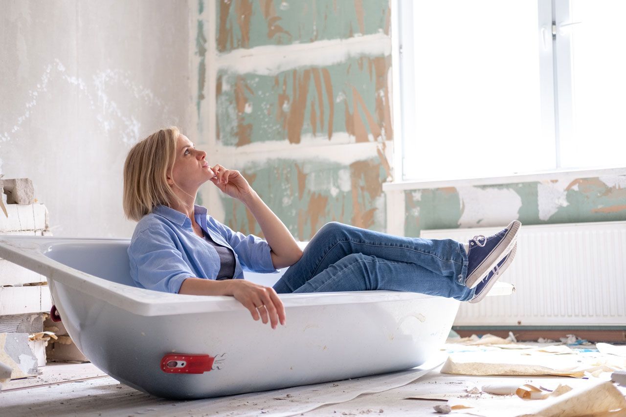 Woman relaxes in a bathtub amidst a construction site, gazing upwards, wearing jeans and a blue shirt.