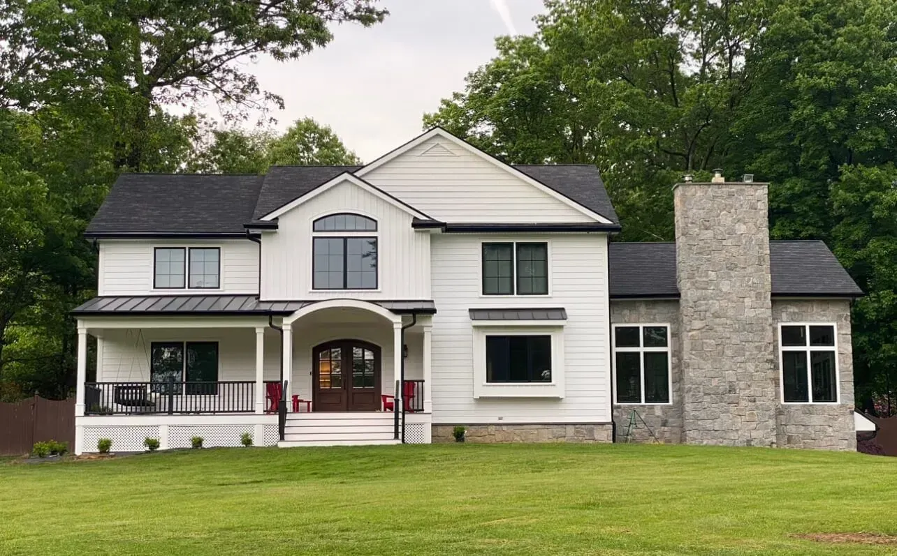 White house with black roof, stone chimney, and front porch. Green grass and trees in the background.