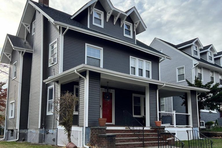 Dark gray two-story house with a red front door, porch, and dormers under a cloudy sky.