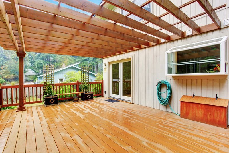 Wooden deck with pergola, French doors, and bay window; hose, storage box, and plants.