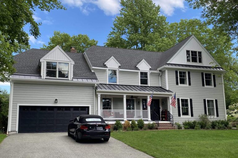 Two-story house with light gray siding, black garage door, car in driveway, blue sky, and green trees.