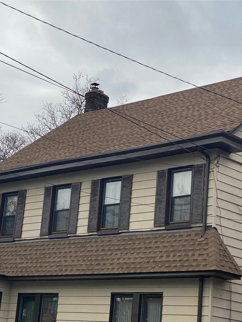 Two-story house with tan siding, brown roof and shutters, chimney with a cap, and power lines against a cloudy sky.