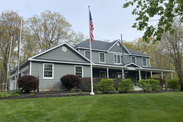 Two-story gray building with a porch, American flag, and surrounding lawn and trees.