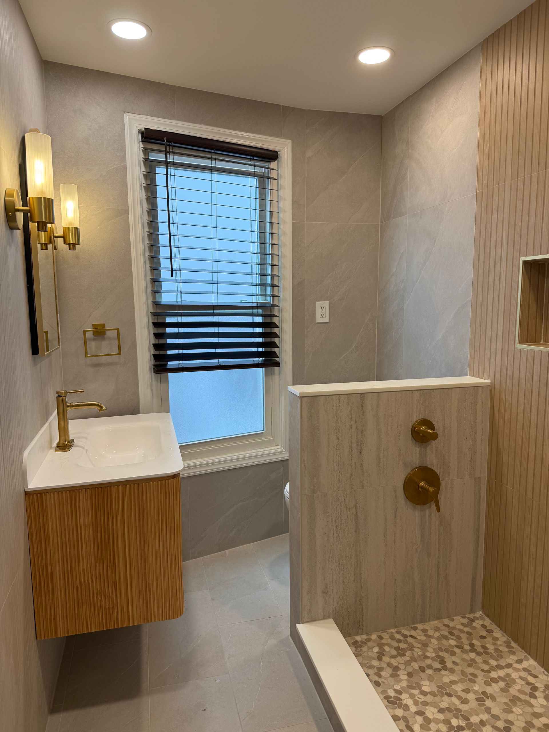 Modern bathroom with walk-in shower, floating vanity, brass fixtures, and window with blinds.