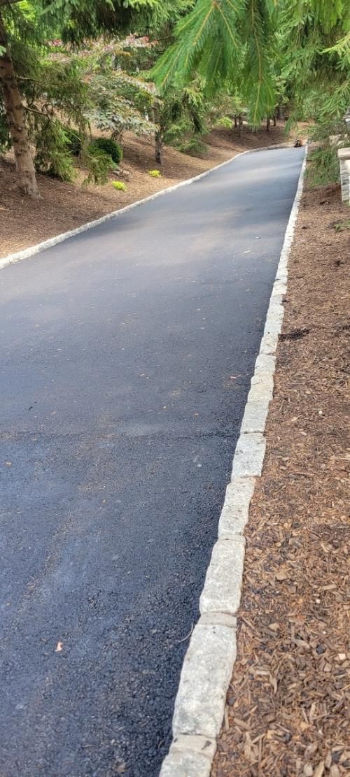 Asphalt driveway bordered by stone and mulch, leading through a wooded area.