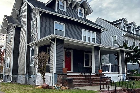 Two-story house with dark gray siding, a red front door, and a porch with brick steps. Overcast sky.