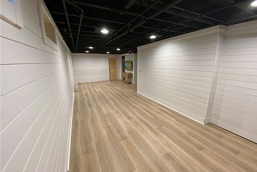 Empty room with white paneled walls, light wood-look floor, black ceiling, and recessed lights.
