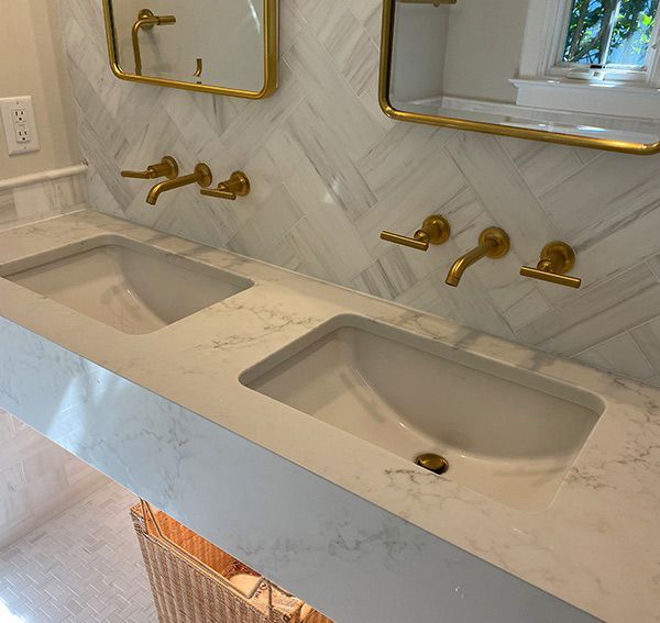 Double bathroom vanity with white countertop, sinks, and gold fixtures, set against a marble tile wall.