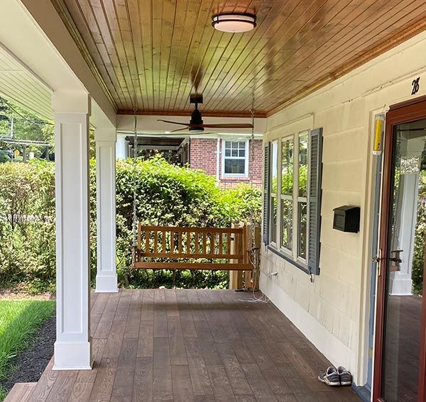Covered front porch with wood ceiling, white columns, ceiling fan, and a bench overlooking a garden built by American Home Remodeling