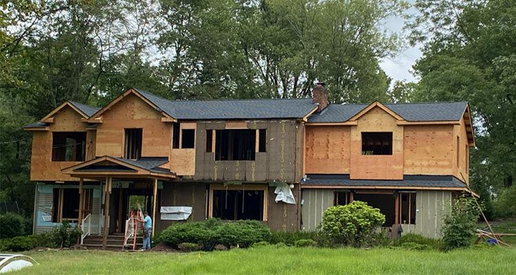 House under construction; unpainted siding, exposed wood, two people on porch, green grass, trees.