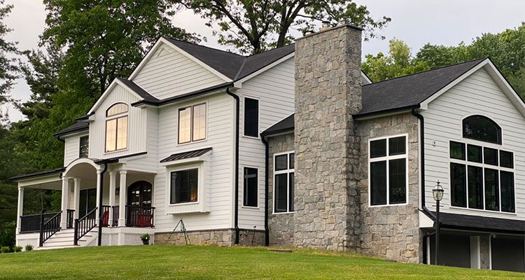 Two-story white house with black roof, stone chimney, and large windows on a grassy hill.