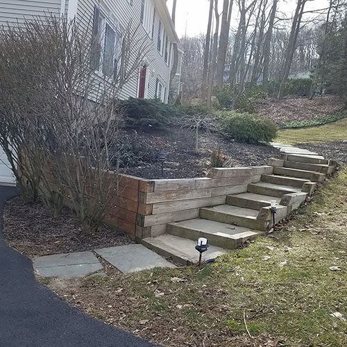 Steps and retaining wall leading to a house, constructed of wood and stone, on a grassy hill.