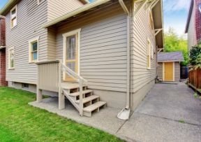 Tan house exterior with stairs leading to a door, concrete path, and a small shed.