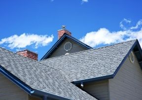 Gray shingled roof with a dark blue trim against a bright blue sky with fluffy white clouds, chimneys.