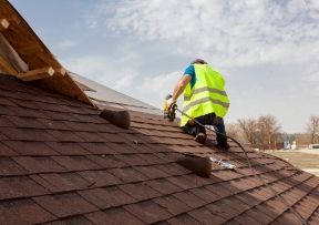 Roofer in safety vest using a nail gun on a brown shingle roof under a partly cloudy sky.