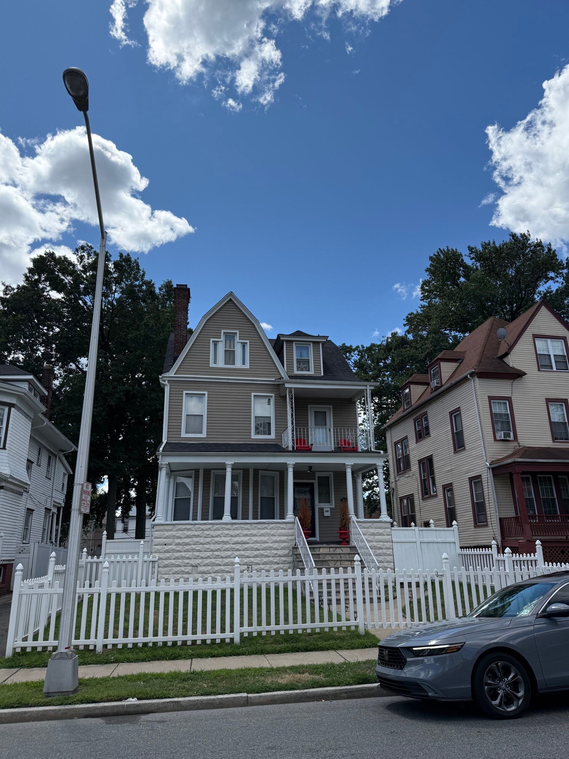 Three-story house with a white picket fence, gray car, and blue sky with clouds.