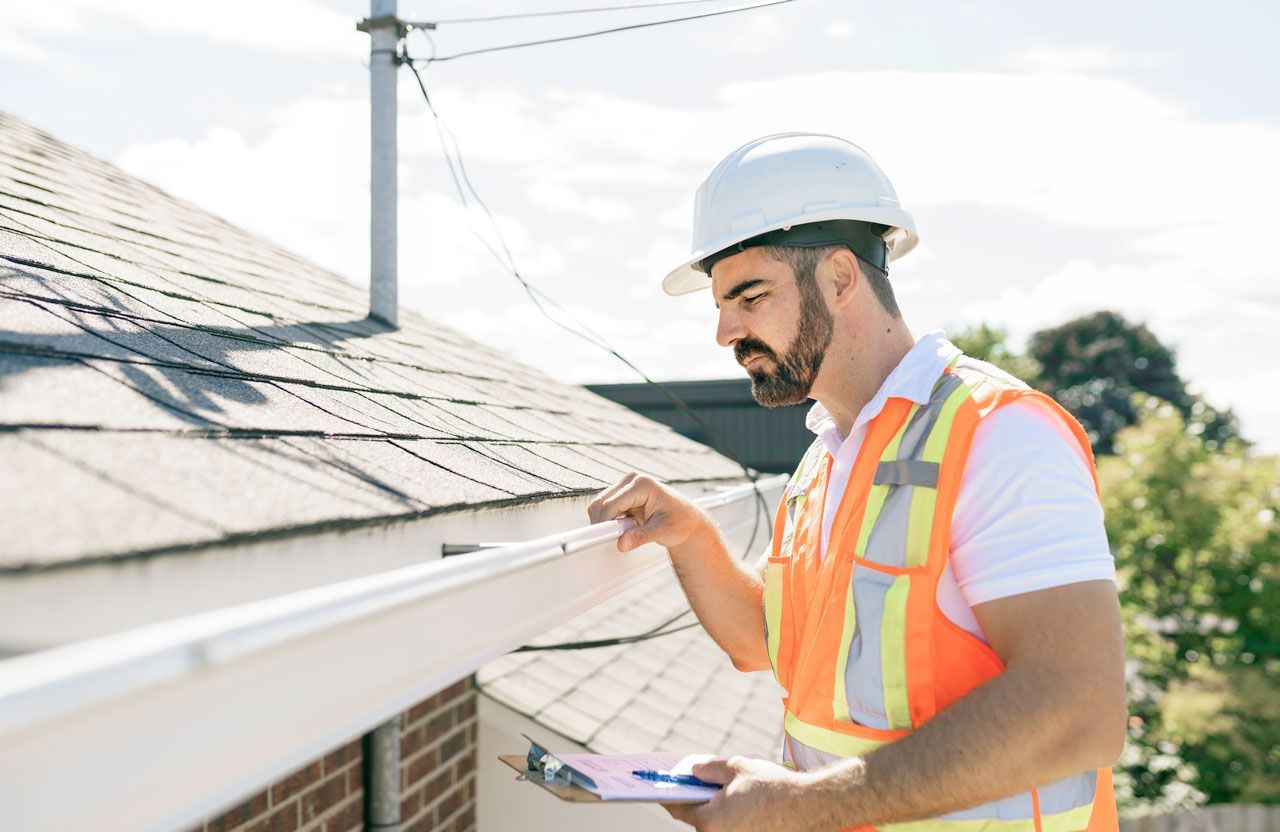 Man in hard hat and safety vest inspecting a roof gutter on a sunny day.