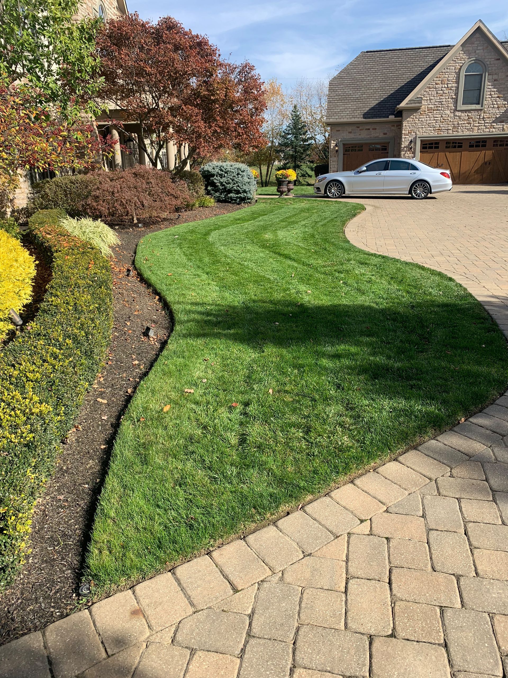 Lush green lawn curving between a brick-paved driveway and garden beds filled with colorful plants.