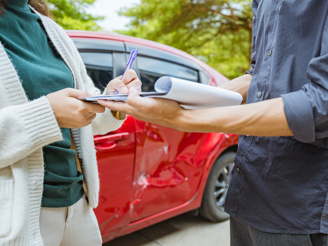 Person writing notes while speaking with another individual beside a damaged red car, assisted by a home and auto insurance broker service in Maricopa, AZ