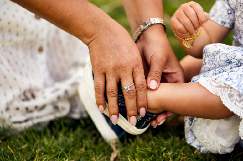 An adult placing a dark blue sneaker on a child's foot while sitting on green grass.