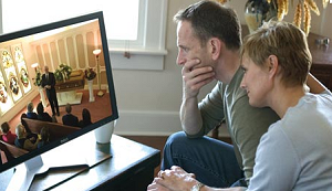 A couple sits together watching a live-streamed funeral service on a computer monitor at home.