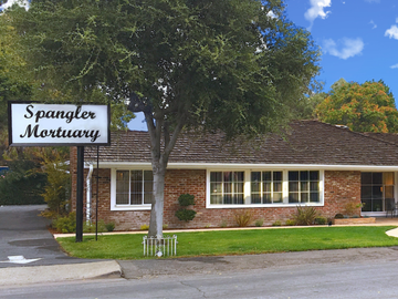 A brick Spangler Mortuary building with a dark wood shingle roof, a sign in the front, a lawn, and a large tree.