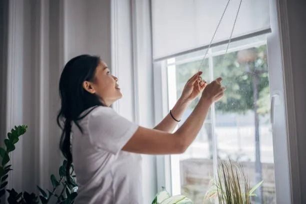 A woman is hanging blinds on a window in a living room.