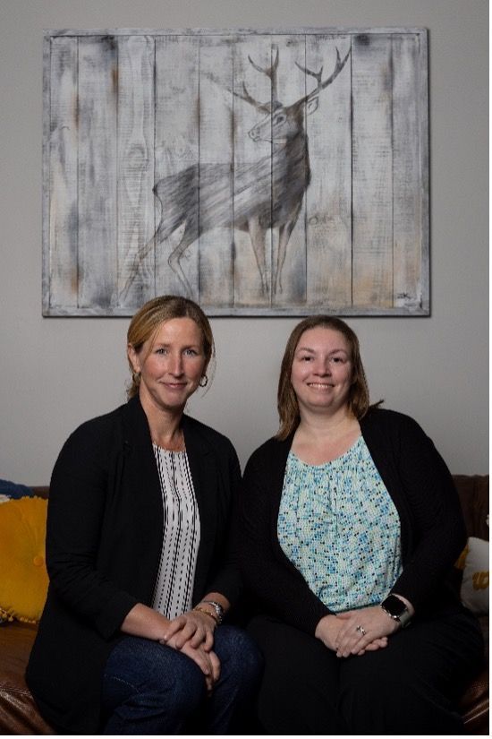 Two women seated indoors beneath a deer painting, smiling at the camera.