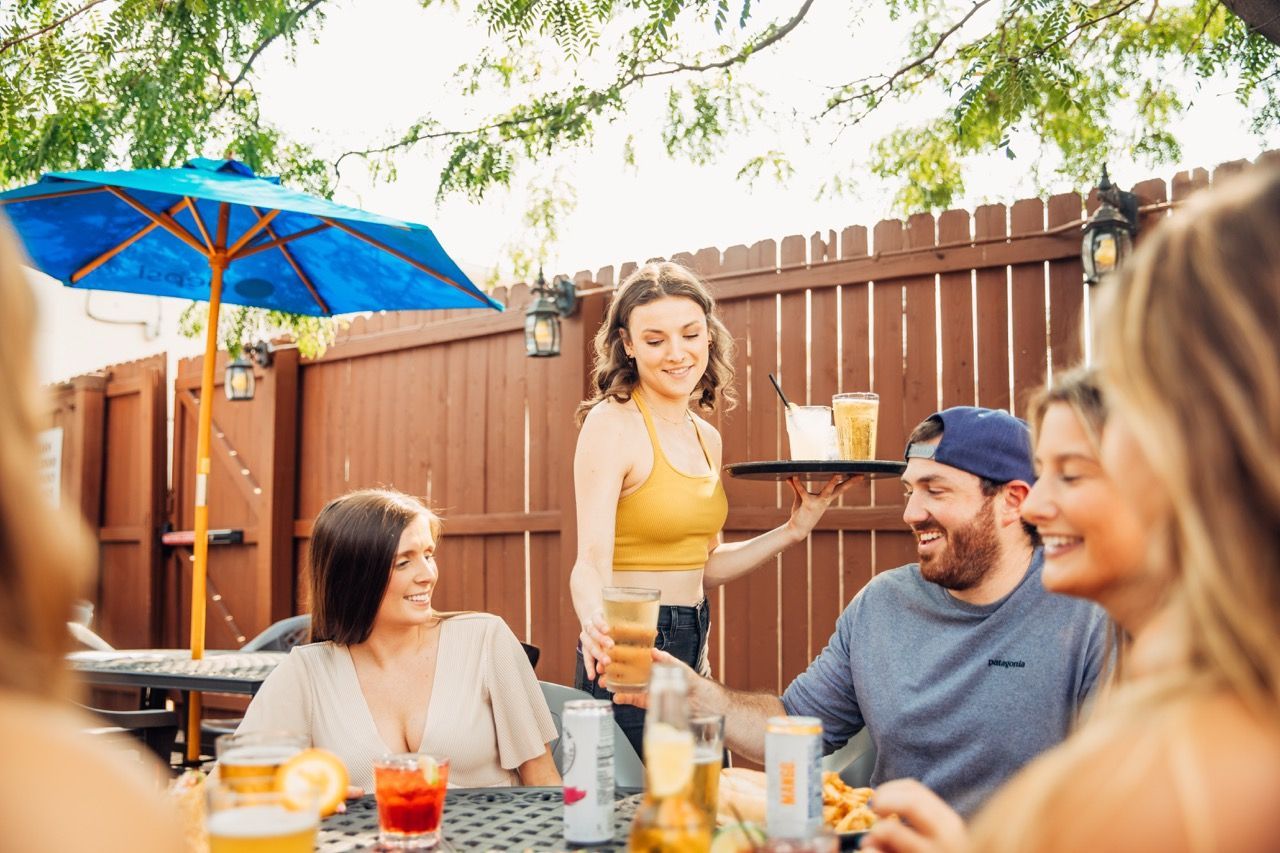 Friends sharing drinks at an outdoor patio table under a blue umbrella, smiling in warm sunlight