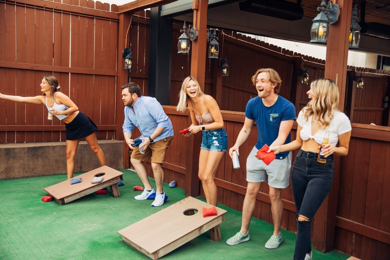 Five people playing cornhole on a green turf patio beside a wooden building