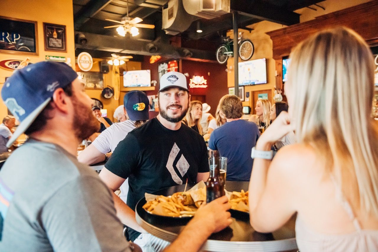 Friends chatting at a lively bar, with one man holding a beer and a plate of chips on the table.
