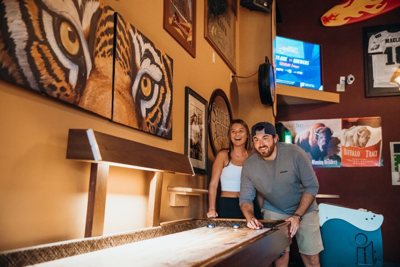 Two people leaning over a pool table in a warmly lit arcade room with tiger wall art and a TV screen.