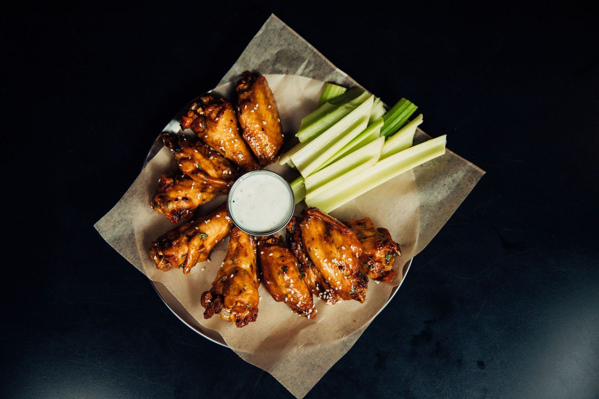 Buffalo wings and celery sticks arranged around a cup of dipping sauce on parchment paper
