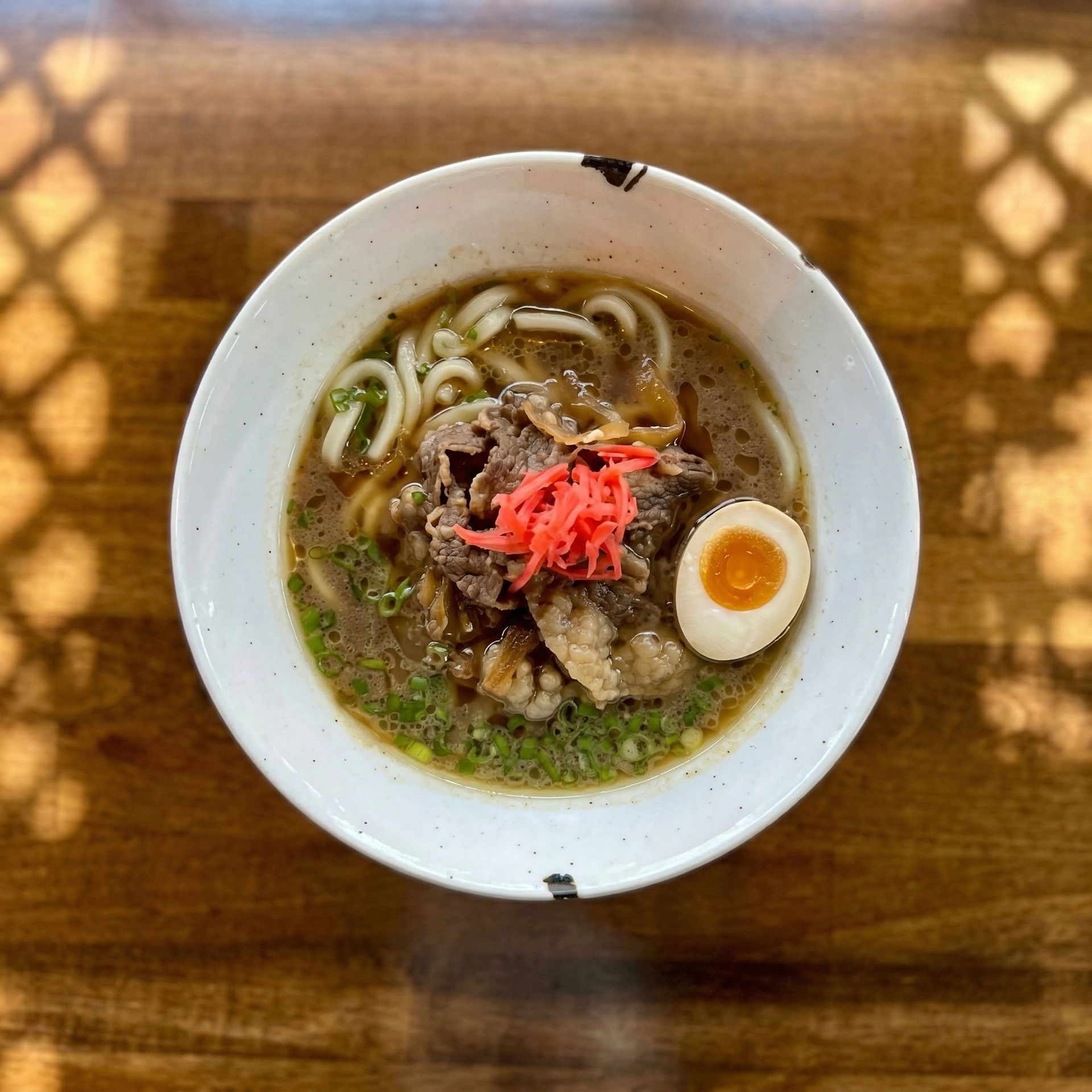 A bowl of udon noodle soup topped with thinly sliced beef, a soft-boiled egg, and pickled ginger on a wooden table.