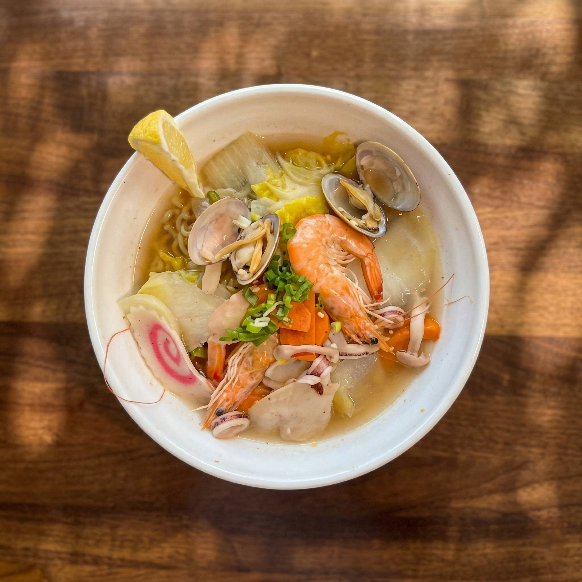 A bowl of seafood ramen with shrimp, clams, narutomaki fish cake, and a lemon wedge, set on a wooden table.