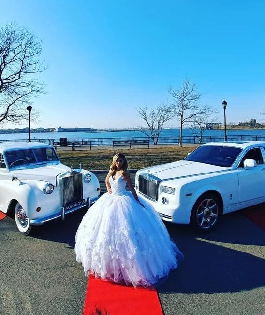 A woman in a wedding dress is standing next to two white cars on a red carpet.