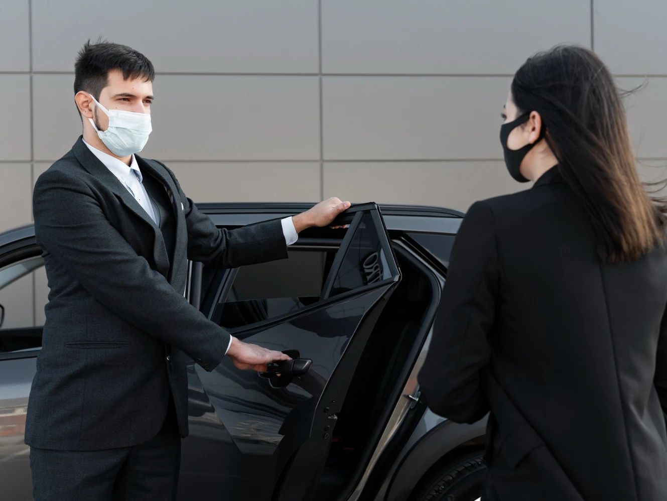 A man wearing a mask is opening the door of a car for a woman.
