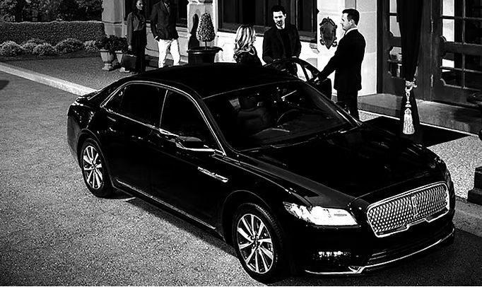 A black and white photo of a man standing next to a black car.
