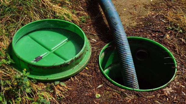 Green septic tank lid open with a blue hose inside, set in the dirt.