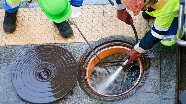 Workers cleaning a manhole with high-pressure water. One worker wearing a green hard hat, the other a red one.