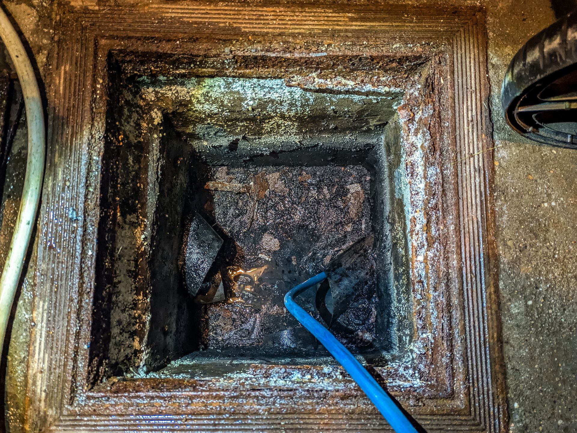 Square drain, dark inside, being probed by a blue tool. Textured frame with encrusted debris.