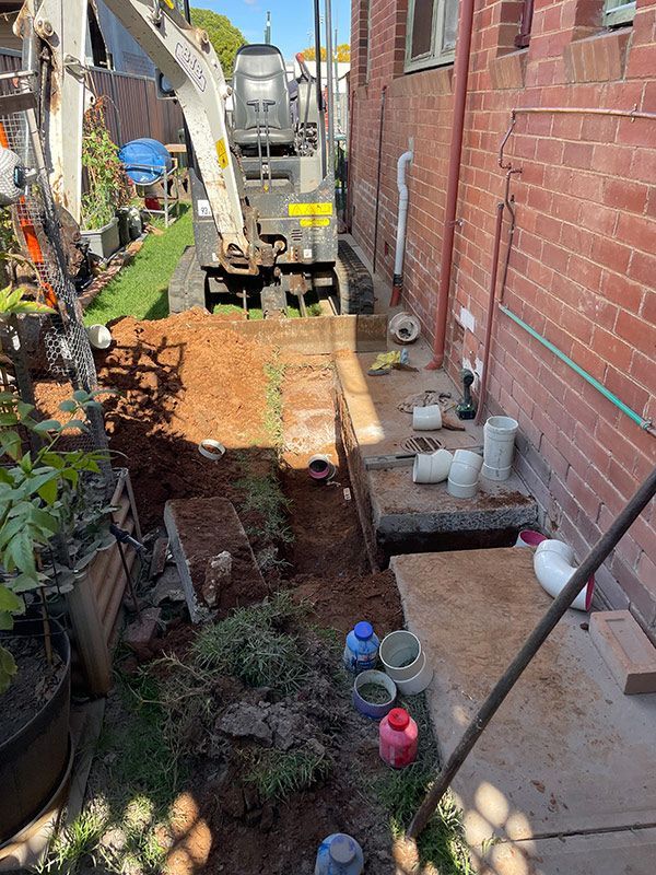 An Excavator Is Digging a Hole in The Ground in Front of A Brick Building — BJB Plumbing & Gasfitting in Coonabarabran, NSW