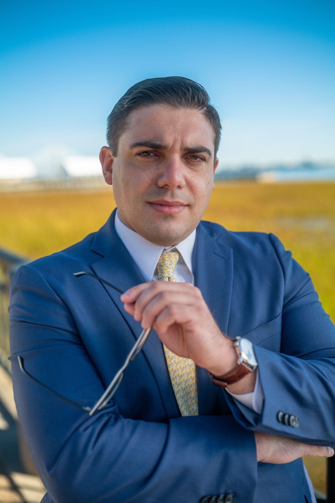 Man in blue suit, holding glasses, outside with a blurred background.