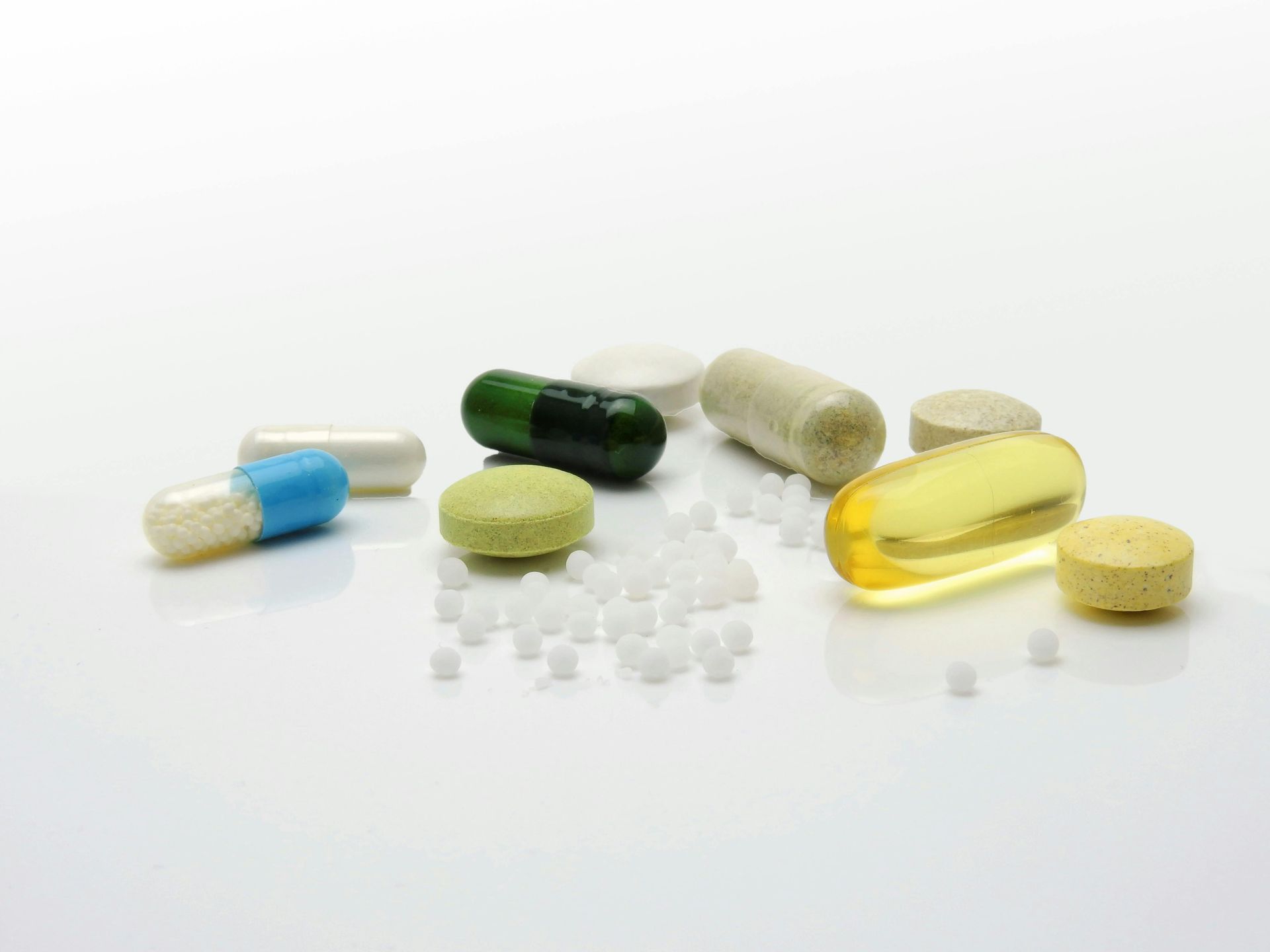 Assorted pills and capsules in various colors on a white surface, with scattered white granules.