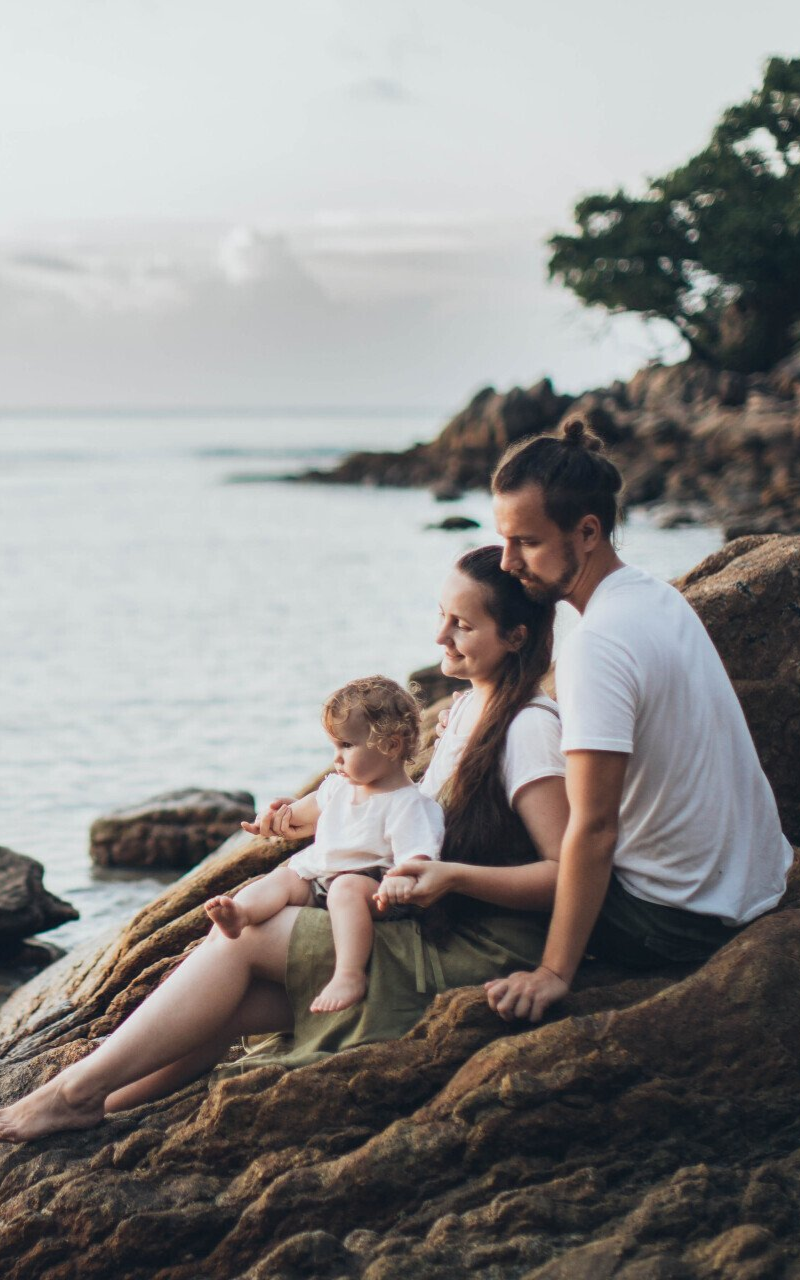 Family of three sitting on rocks by the ocean; father hugs mother, holding child.