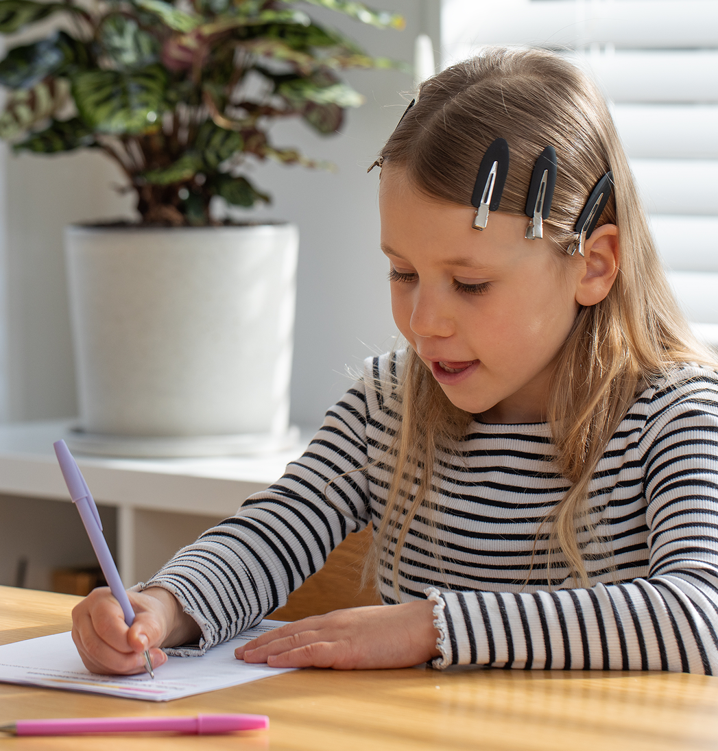 Child writing with a pencil at a table beside a potted plant, wearing a striped shirt and hair clips.