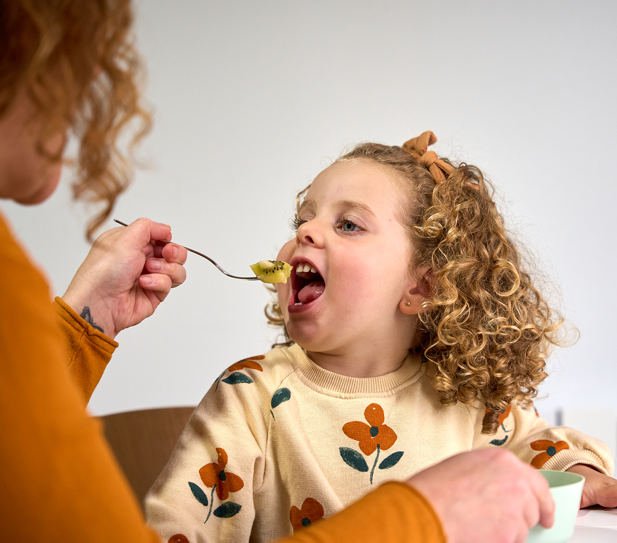 Adult feeding a toddler with a spoon at a table, both in warm-toned clothing.