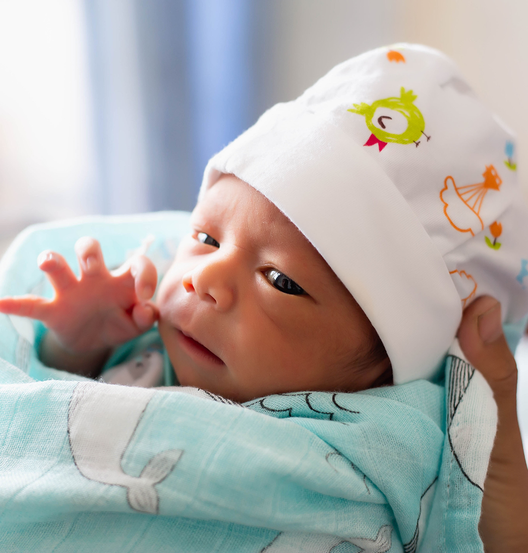 Newborn wrapped in a mint blanket, wearing a white hat, looking upward with a tiny hand raised
