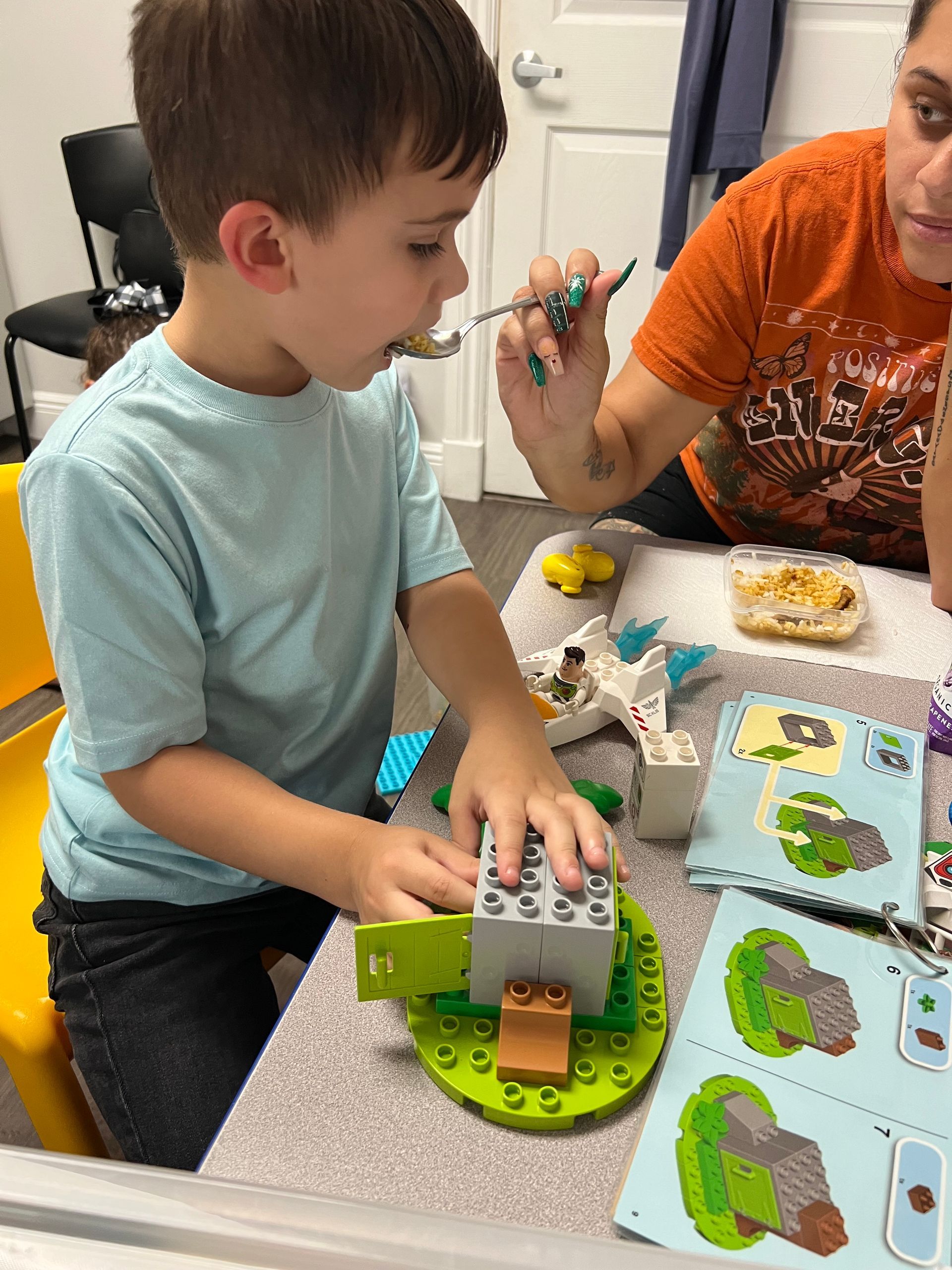 Child opening a green toy set at a table while an adult points beside a snack plate and activity cards.