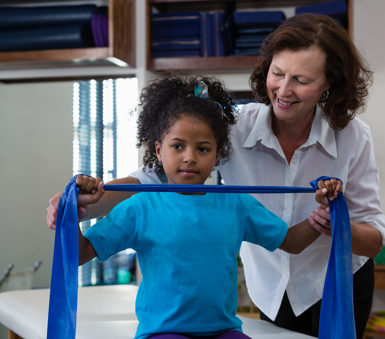 Woman assisting child stretching a blue resistance band in a bright room